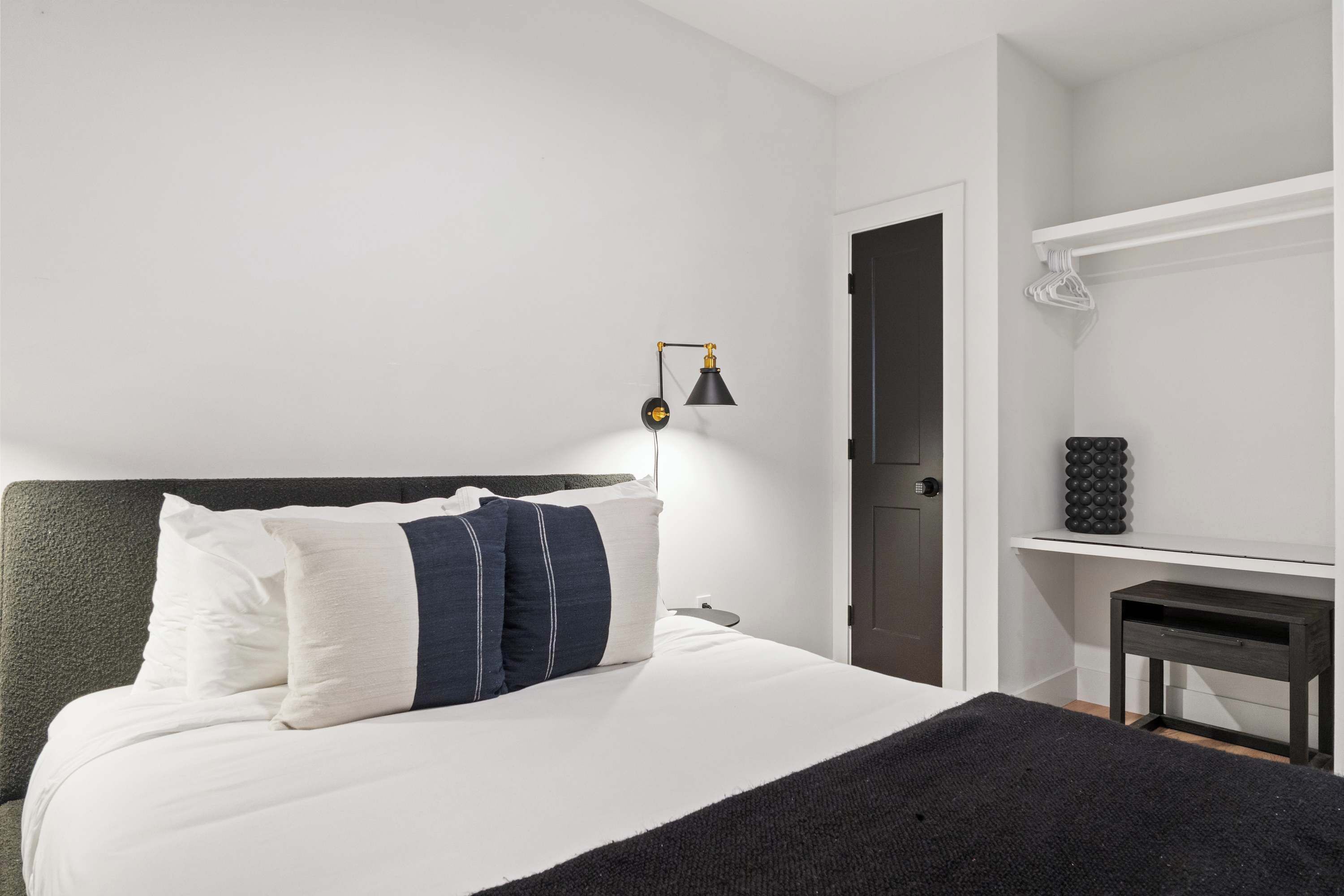 A neatly made bed with pillows, a bedside lamp, a door, and a small table with a decorative item.