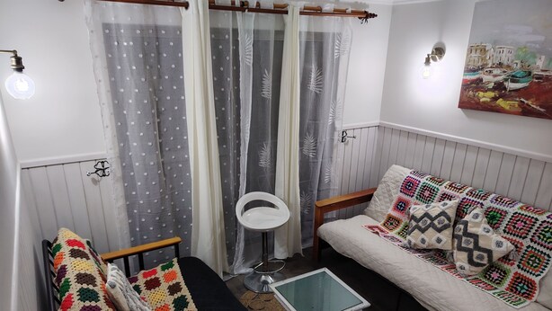 Una habitación acogedora con una cama, una mesita pequeña y una silla. Se ve una ventana con cortinas, una lámpara de pared y un cuadro.