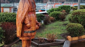 Una estatua de madera de una figura vestida con atuendo tradicional, ubicada en un área ajardinada con vista a un puerto deportivo y edificios al fondo.