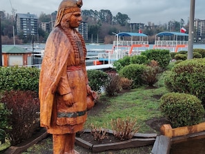 Eine hölzerne Statue einer Person in traditioneller Kleidung, vor einer gepflegten Anlage mit Blick auf einen Yachthafen und Gebäude im Hintergrund.