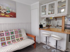 Ein gemütliches Zimmer mit einem weißen Sofa, einer bunten Häkeldecke, einer Holztheke mit Hockern und einer Küchenzeile mit Schränken und einem Kühlschrank.