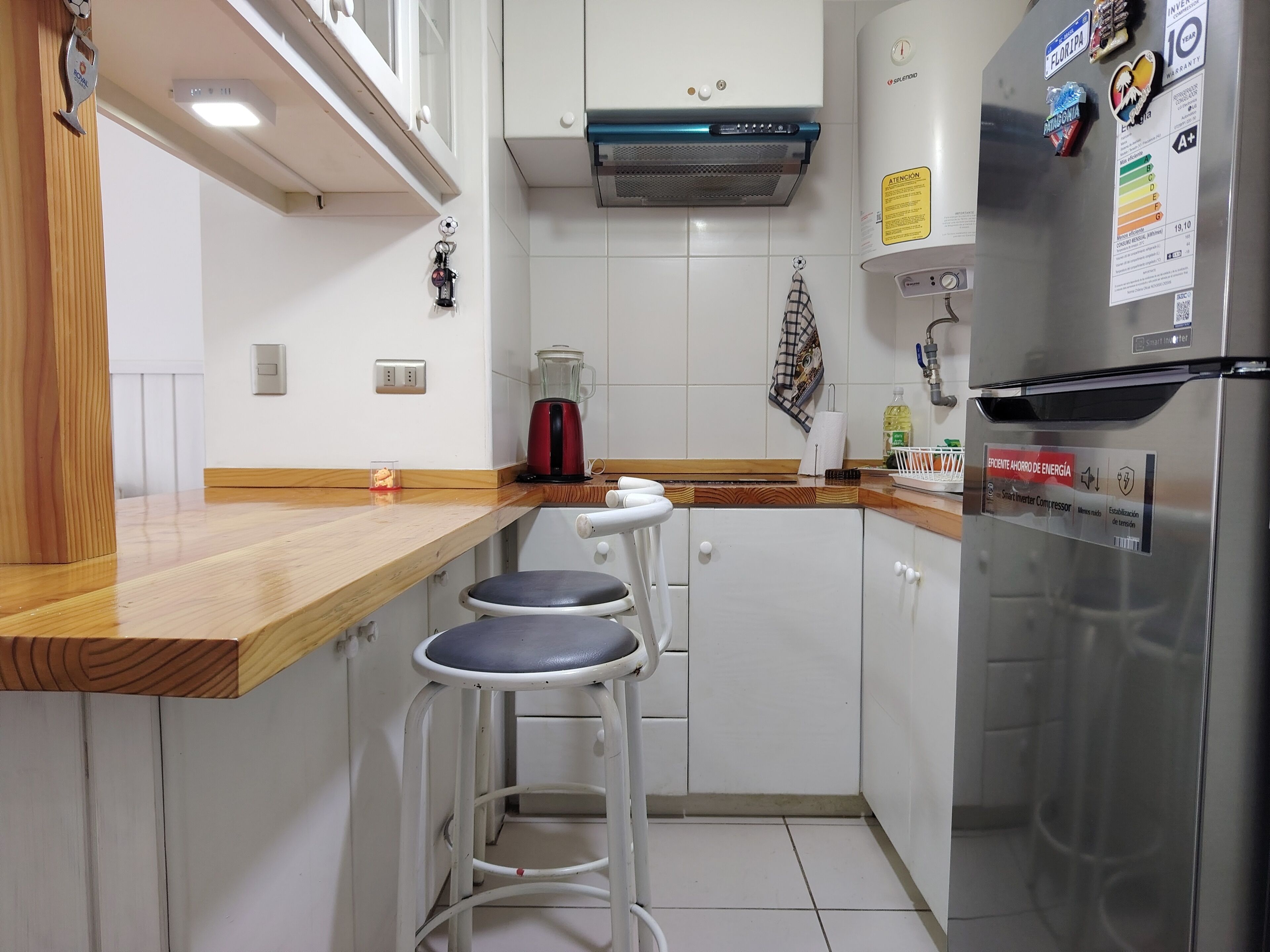 A kitchen with a stainless steel refrigerator, a coffee maker, and a red kettle. There are two white bar stools and a wooden countertop.