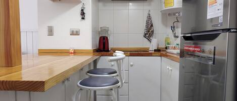A kitchen with a stainless steel refrigerator, a coffee maker, and a red kettle. There are two white bar stools and a wooden countertop.