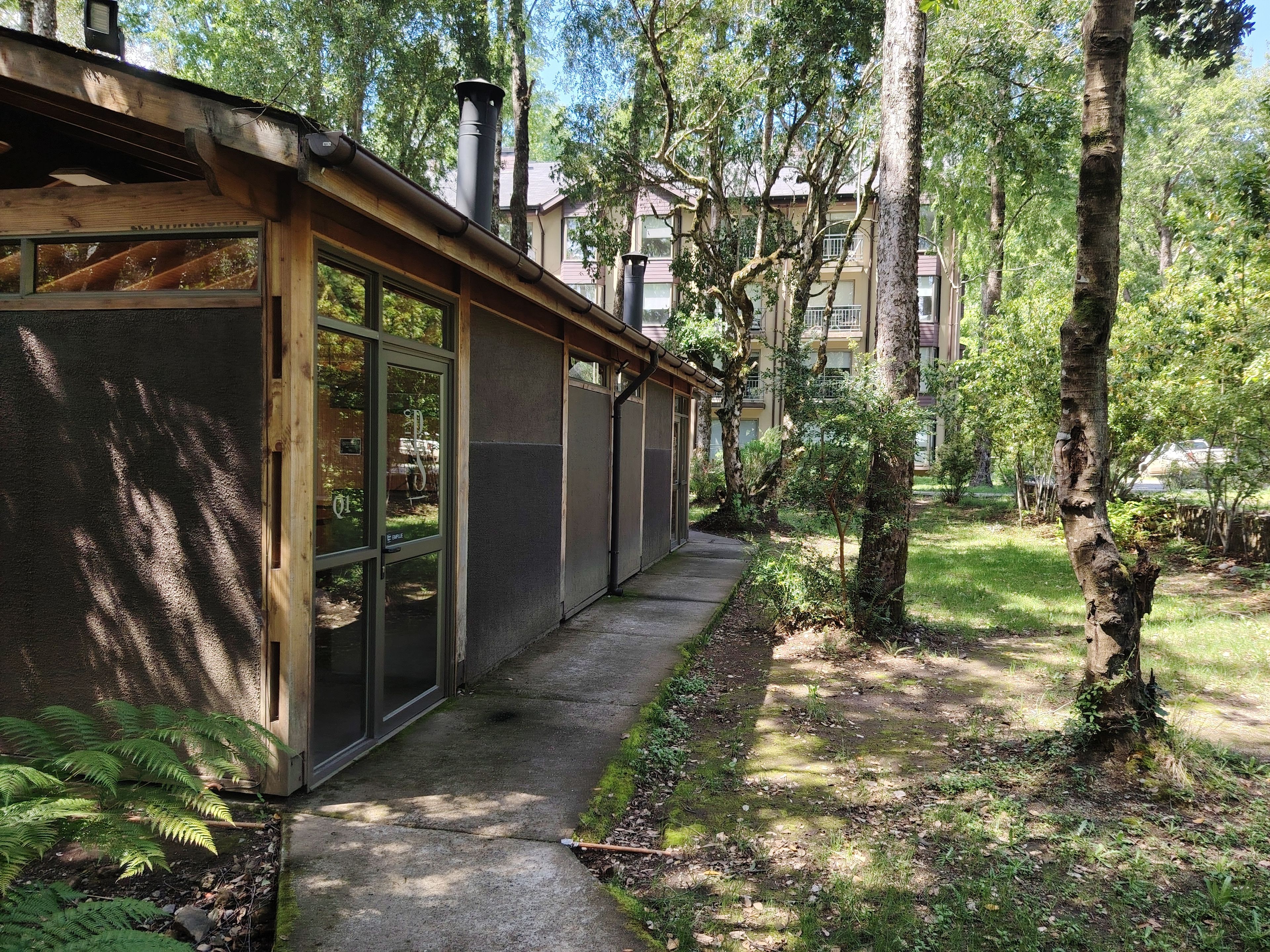 A modern building with large windows, a concrete pathway, and a forested area.