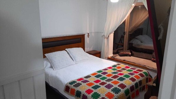 A single bed with a white duvet and a colorful crocheted blanket, a bedside table, and a mirror with a wooden frame. - A warm and cozy apartment located in a peaceful, natural, and relaxed setting (Araucanía)