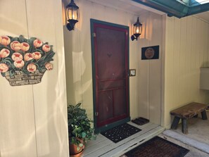 En trädör med rød ramme, en velkomst måtte, en bænk og en potteplante på verandaen.