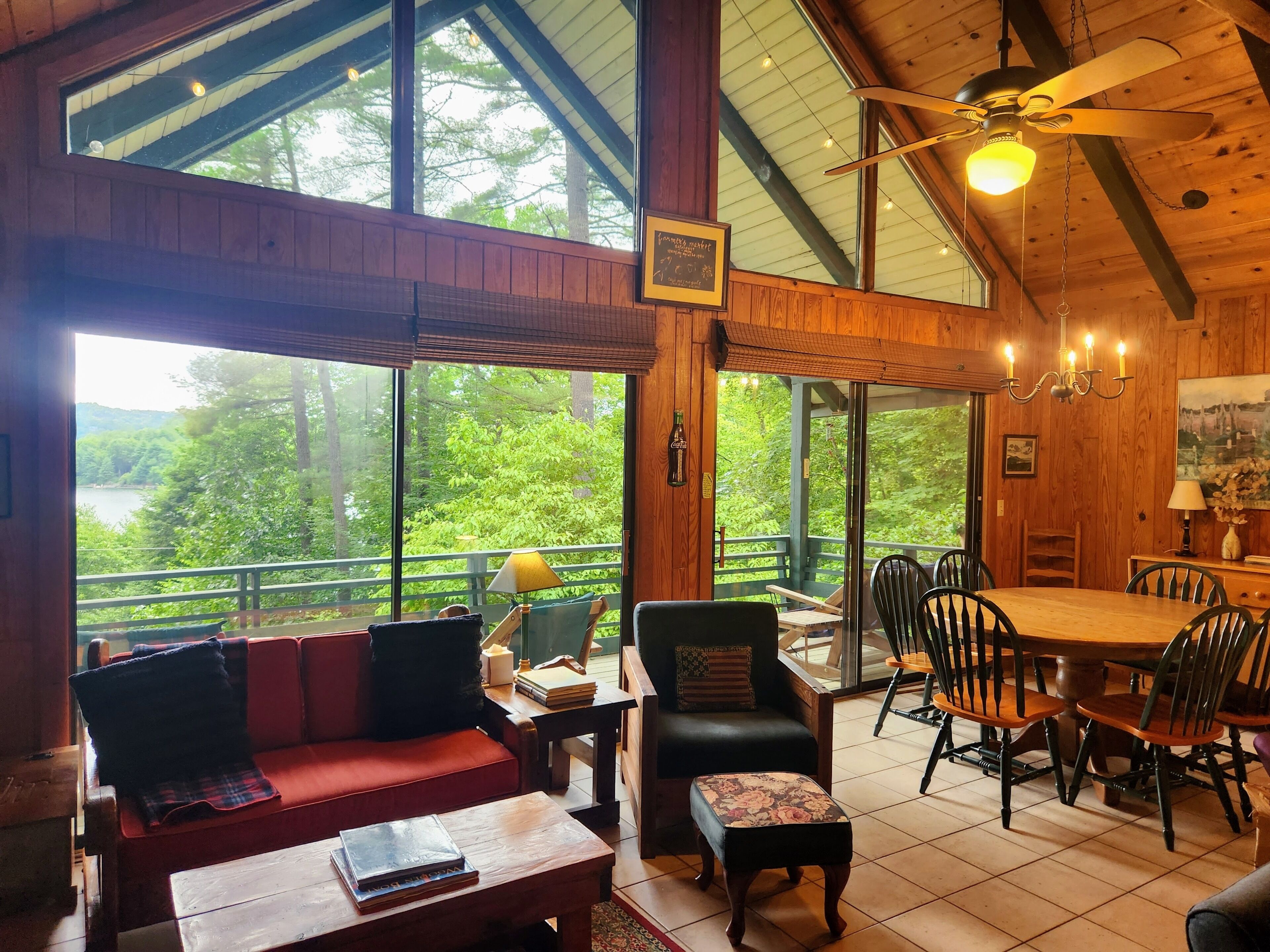 A spacious room with a large wooden ceiling, a dining area with a table and chairs, a sofa, and a view of a forest through large windows.