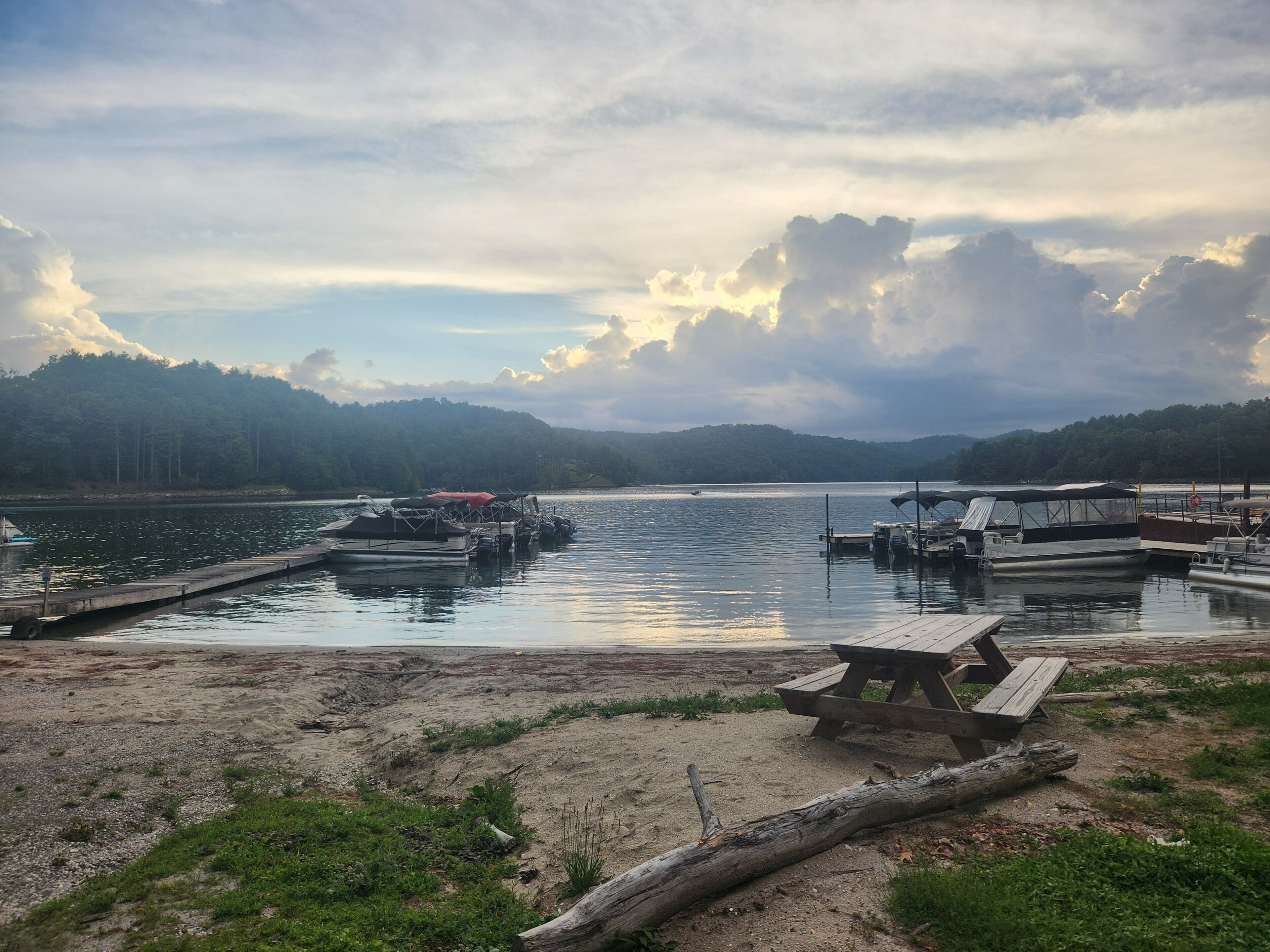 A lakeside dock with boats, a picnic table, and a forested hill in the background.