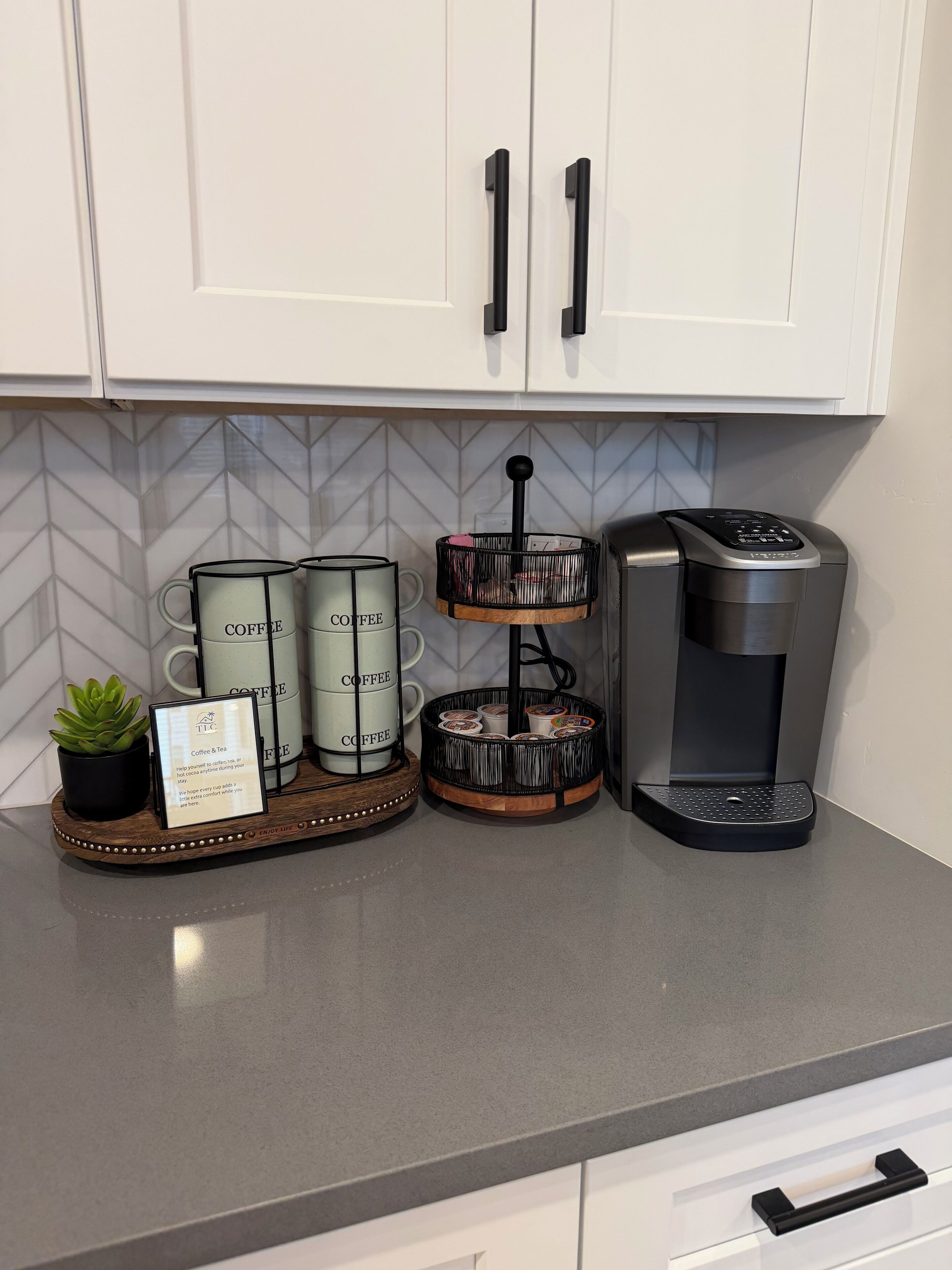 A kitchen counter with a coffee maker, two coffee mugs, a tray with a plant, and a sign.