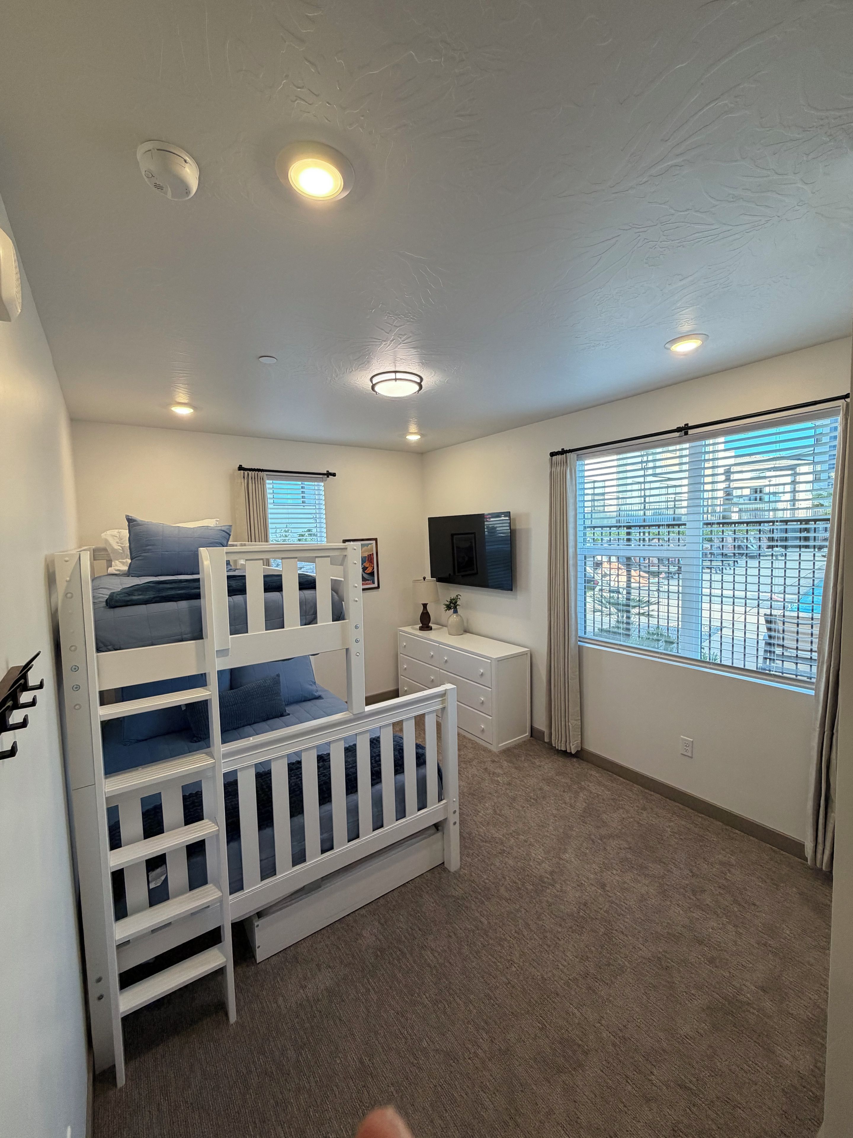 A child's bunk bed with a TV above it, a bedside table, and a window with blinds.