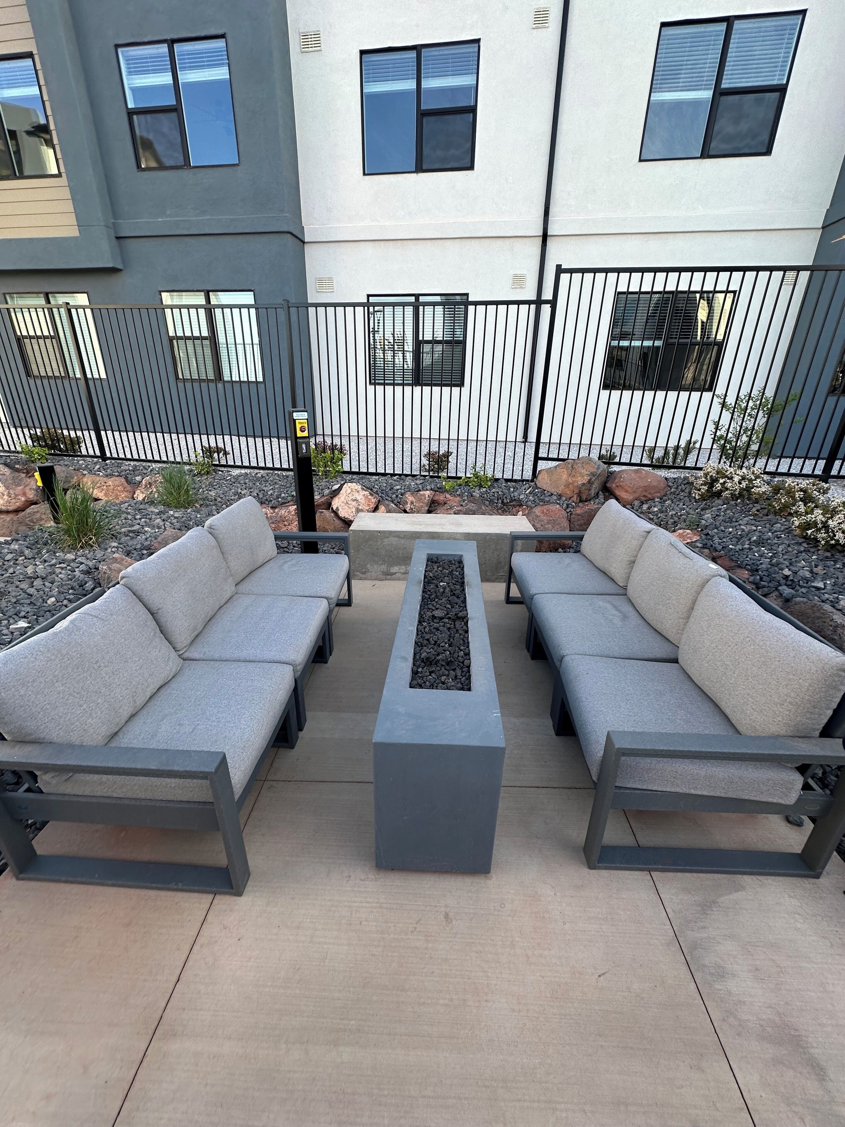 An outdoor seating area with two sofas and a central table, surrounded by a metal fence and a building with windows.