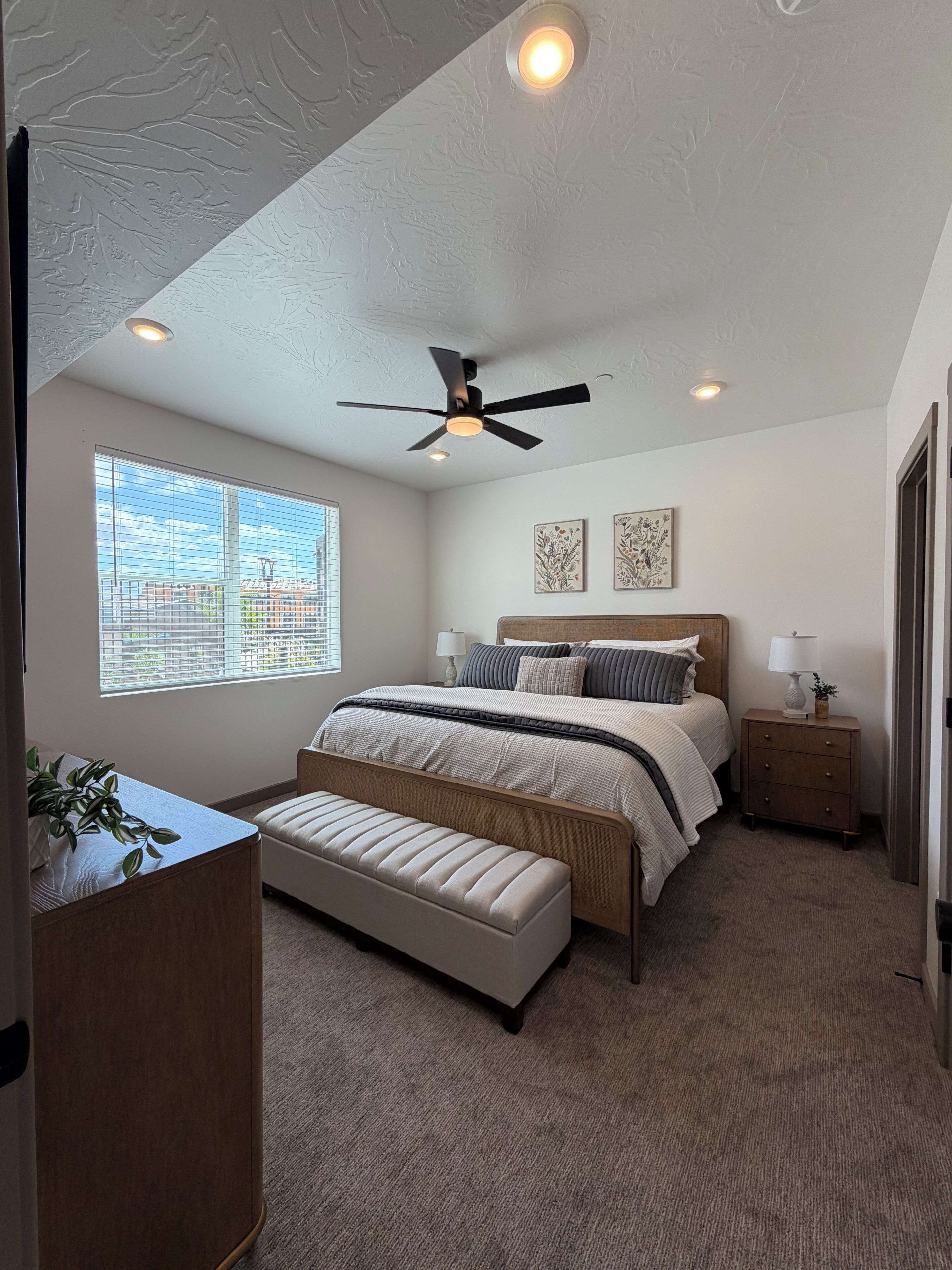 A bedroom with a bed, a ceiling fan, a window with blinds, a bedside table with a lamp, and a plant.
