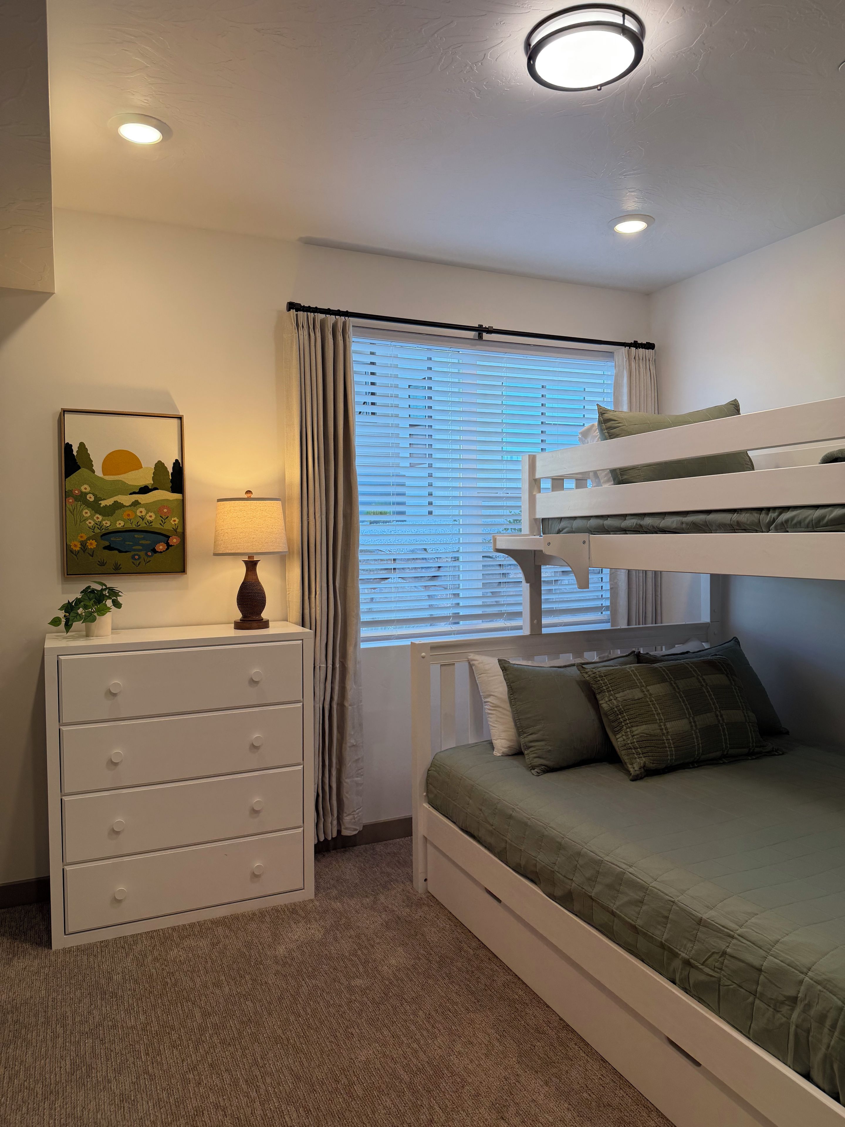 Un lit superposé avec un matelas vert, une commode blanche avec des tiroirs, une lampe et une fenêtre avec des stores.