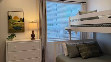Un lit superposé avec un matelas vert, une commode blanche avec des tiroirs, une lampe et une fenêtre avec des stores.