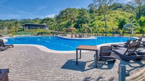 Ein Swimmingpool mit Liegestühlen und einem Tisch, umgeben von Grünpflanzen und einem Gebäude im Hintergrund.