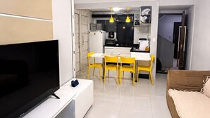 Uma sala de estar moderna com televisão de tela plana, um sofá e um espaço de jantar com cadeiras amarelas e uma mesa branca.