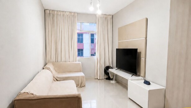 Quarto de hotel moderno com sofá, televisão, mesa pequena e janela com cortinas.