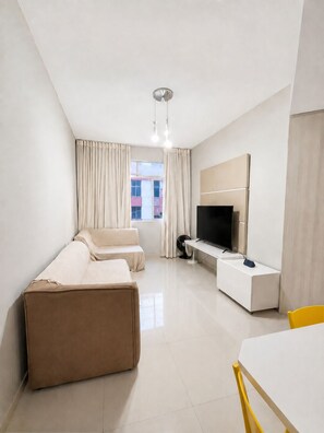 Una habitación de hotel moderna con sofá, televisión, una mesita y ventana con cortinas.
