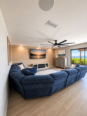 A modern living room with a sectional sofa, a flat-screen TV mounted on a wooden wall, a ceiling fan, and a large window offering a view of the outdoors. - Blue Horizon • 5 minutes to Launch Ramp + Lake Views + Game Room! (Lake Havasu City)