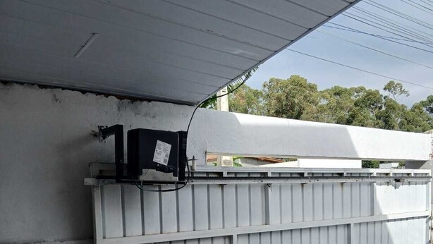 A garage with a white car parked inside, a metal fence, and a black electrical box mounted on the wall. - House 5 minutes from the Mozambique dunes trail (Florianópolis)