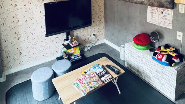 Habitación con una televisión de pantalla plana montada en la pared, una pequeña mesa de madera con revistas, dos otomanes grises, una obra de arte con un mapa del mundo y un gorro rojo en el suelo.