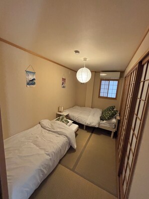 A small, narrow room with two single beds, a hanging light fixture, and a window with a view. - Straight from KIX! Easy Access to Kyoto & Nara / Spacious House for Families & Groups (大阪市西成区千本北)