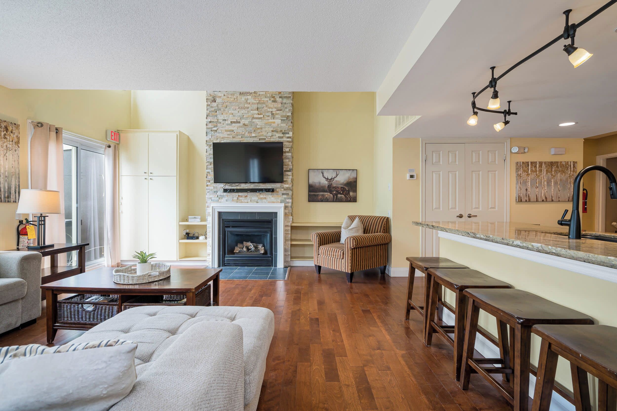 A spacious living area with a fireplace, a television mounted on a stone wall, a comfortable armchair, and a kitchen area with a bar counter.