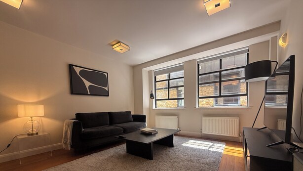 Una sala de estar moderna con sofá, mesa de centro y mueble para televisor. Hay ventanas amplias con paredes de ladrillo y un radiador blanco.