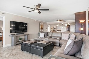 Ein modernes Wohnzimmer mit einem Ecksofa, einem Flachbildfernseher, einem Deckenventilator und einem Essbereich im Hintergrund.