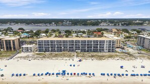 Un hotel frente a la playa con varios pisos, una playa arenosa al frente y vistas al agua y al paisaje urbano de fondo.