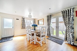 A dining area with a wooden table and white chairs, a vase with flowers, and a view of the outdoors through a glass door. - Lodge 4 - Pavilion (Brigg)