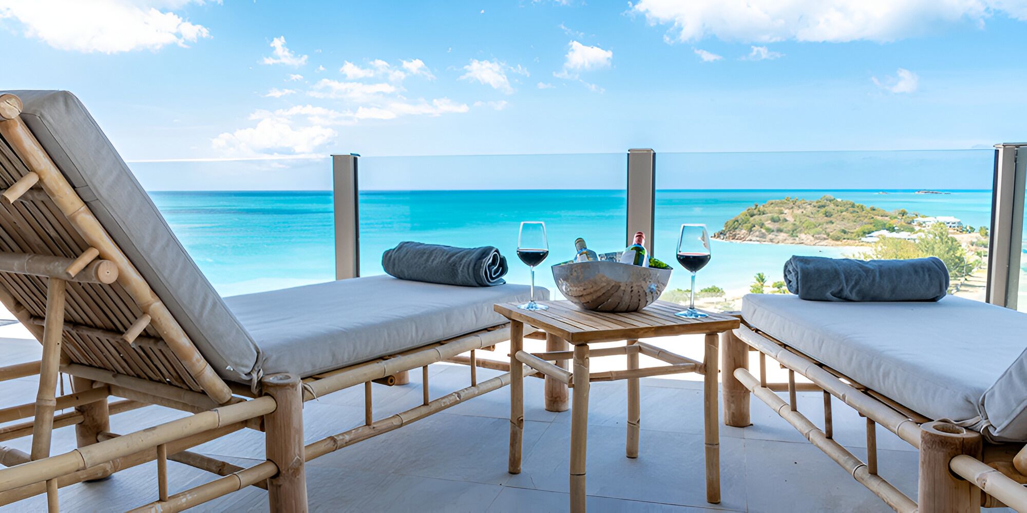 A wooden deck with two lounge chairs and a table, overlooking a clear blue sea and a distant island.