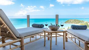A wooden deck with two lounge chairs and a table, overlooking a clear blue sea and a distant island.
