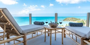 Una terraza de madera con dos sillones y una mesa, con vista a un mar azul cristalino y una isla lejana.
