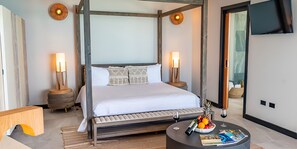 A modern hotel room with a four-poster bed, two bedside tables with lamps, a round coffee table with magazines and fruit, and a large mirror on the wall. - Spacious Antigua Suite with Infinity Pool & Wraparound Terrace (Urlings)