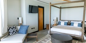A modern hotel room with a four-poster bed, a sofa with patterned cushions, a flat-screen TV mounted on the wall, and a wooden door. - Panorama: Luxury 2-Bedroom Ocean-View Suite with Private Infinity Pool, Antigua (Urlings)