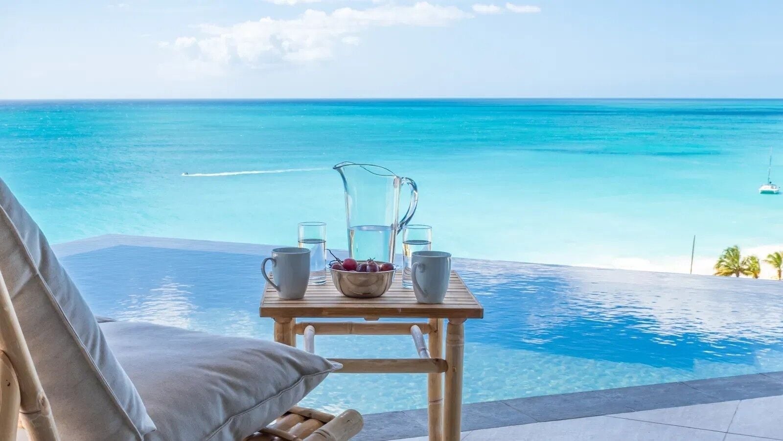 A wooden table with a bowl of fruit, glasses, and a pitcher, overlooking a clear blue ocean and a sandy beach.