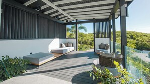 A modern outdoor deck with a wooden floor, two seating areas with cushions, and a glass railing overlooking greenery. - Seastar: Luxury 2-Bedroom Ocean-View Suite with Private Pool, Antigua (Urlings)