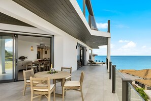 Un área de comedor exterior moderna con vista al mar, que presenta una mesa redonda y sillas, una puerta de cristal que da al interior, y un balcón con barandilla frente al océano.