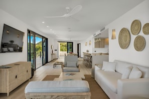 Una sala moderna con sofá, sillón y mesa de centro. Hay un comedor con mesa y sillas. La habitación tiene ventanas amplias y un ventilador de techo.