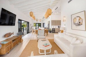 Una sala de estar moderna con un sofá blanco, una mesa de centro y un televisor montado en la pared. Hay una zona de comedor con una mesa y sillas, y una cocina al fondo.
