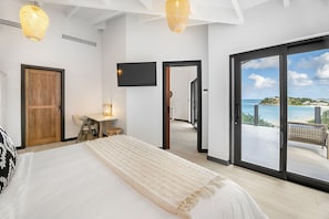 Un dormitorio con una cama grande, un escritorio, una silla y un televisor. Hay una puerta corrediza de vidrio que da a un balcón con vistas al mar.