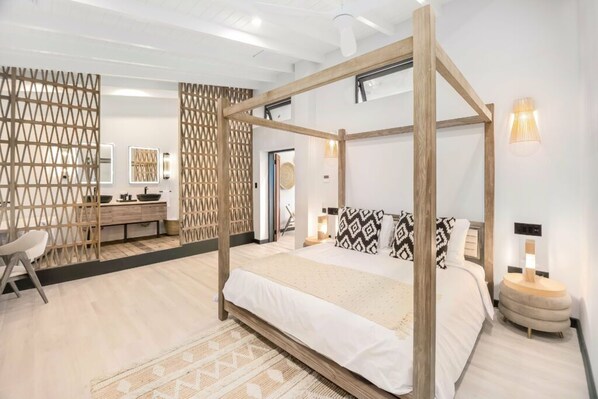 Una cama con dosel, con una colcha blanca y almohadas estampadas, una mesita de noche de madera con una lámpara, y un baño moderno con tocador de madera y un espejo.