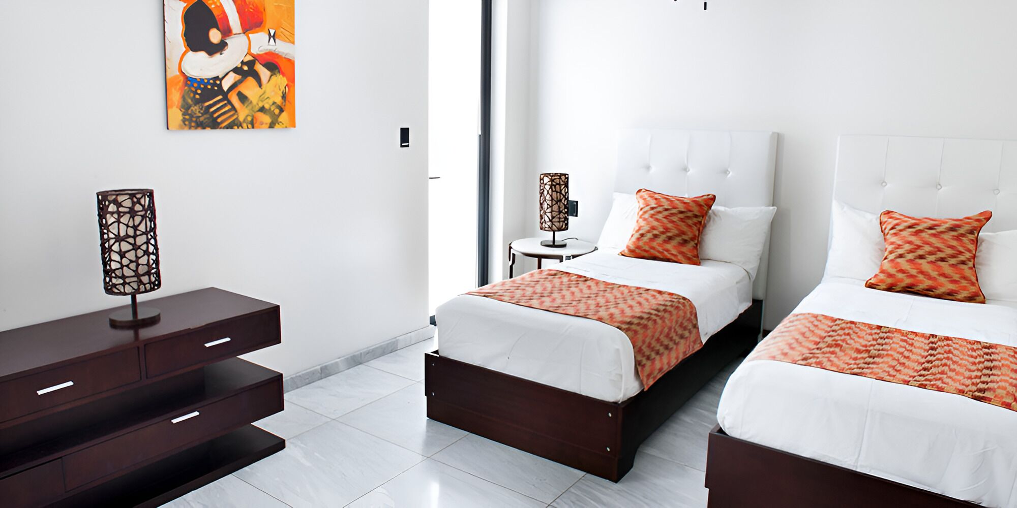 A modern hotel room with two beds, a nightstand, a lamp, and a painting on the wall.