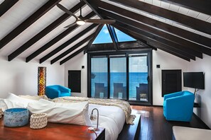 A bedroom with a large bed, a wooden ceiling, a view of the sea, and a TV on the wall.