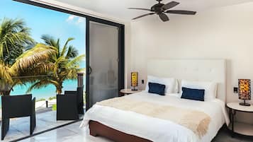 A bedroom with a large bed, two bedside tables, a ceiling fan, and a view of palm trees and the ocean.