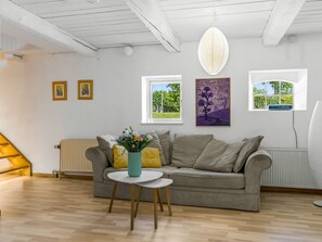 Una sala de estar moderna con un sofá gris, una mesita redonda y un jarrón de flores. Hay dos cuadros en la pared, una ventana con vistas a zonas verdes y un techo blanco con vigas a la vista.