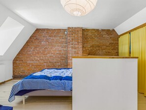 Ein Schlafzimmer mit schrägen Wänden, einer Ziegelwand, einem Bett mit blauer Karobettdecke und einer weißen Kommode.