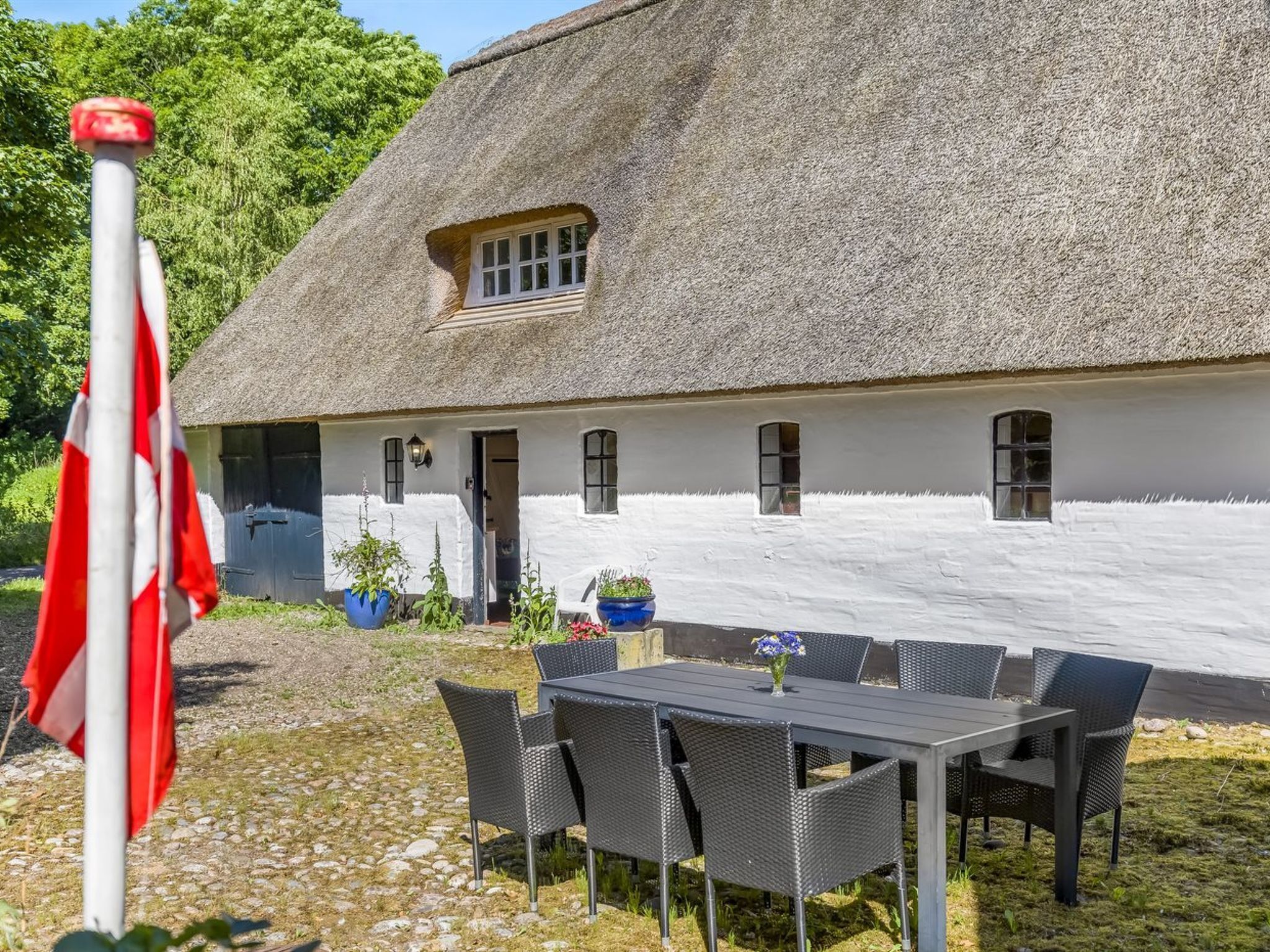 Ein Gebäude mit einem Strohdach und weißer Fassade, ein Essbereich mit Gartenmöbeln und eine Fahnenstange mit einer Flagge.