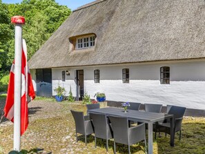 Ein Gebäude mit einem Strohdach und weißer Fassade, ein Essbereich mit Gartenmöbeln und eine Fahnenstange mit einer Flagge.